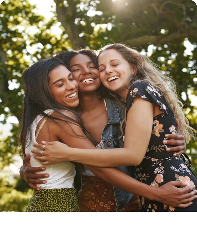 Três mulheres sorrindo e se abraçando em um dia ensolarado, rodeadas por árvores verdes. Amor e amizade em destaque.